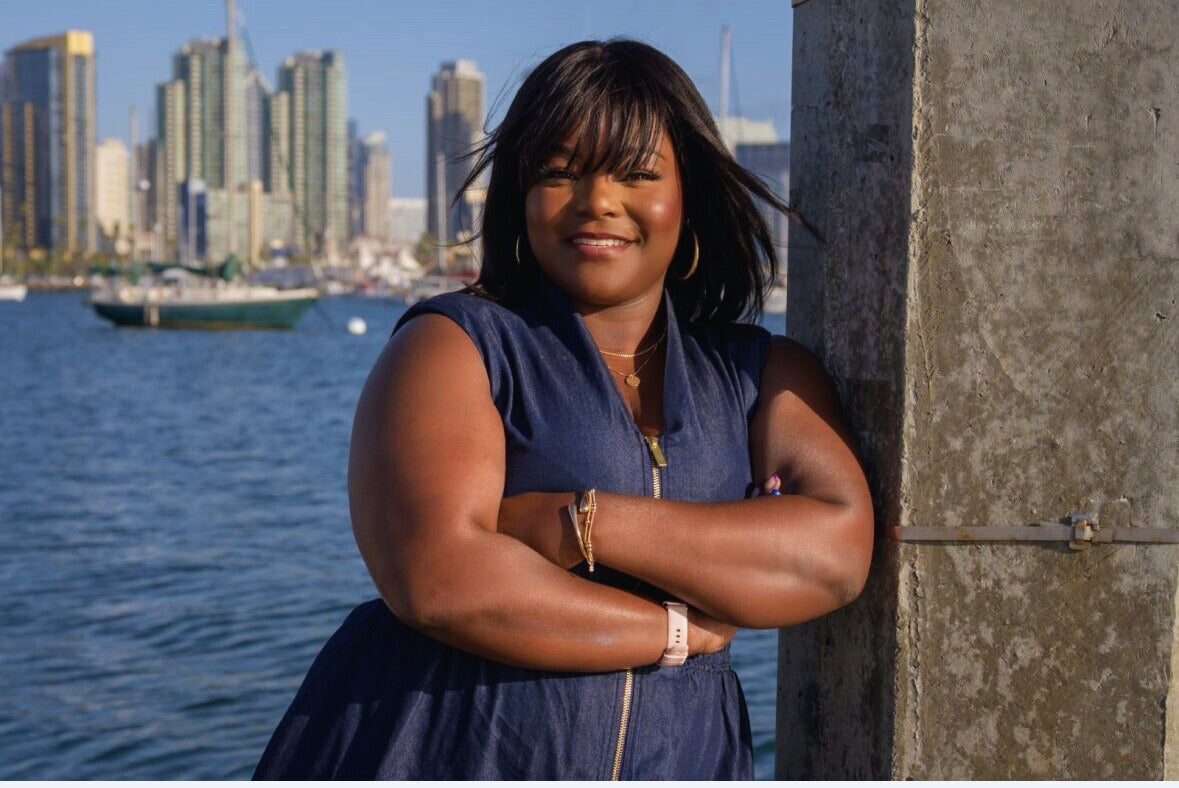 A confident Black woman in a navy dress standing by the water in front of a city skyline, arms crossed and smiling, radiating grounded strength, empowerment, leadership, and soulful confidence.